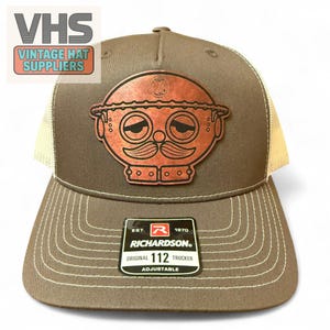 May include: A brown and tan trucker hat with a leather patch featuring a cartoon face with a mustache. The hat has a curved brim and white stitching. The Richardson brand label is visible.