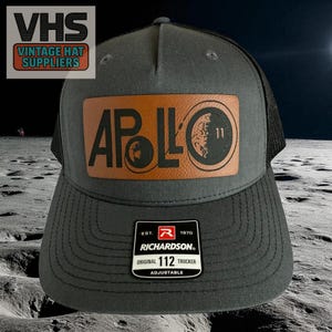 May include: A gray and black trucker hat with a brown leather patch featuring the word "APOLLO" and a graphic of the moon. The hat has a curved brim and a mesh back. The Richardson brand label is visible. The hat is adjustable.