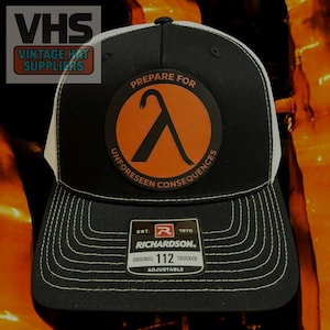 May include: Black and white trucker hat with a patch featuring an orange lambda symbol inside a circle, with the text "PREPARE FOR UNFORESEEN CONSEQUENCES". The hat has a white mesh back and a curved brim with white stitching. The tag reads "RICHARDSON ORIGINAL 112 TRUCKER".