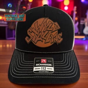 May include: Black and white trucker hat with a brown leather patch featuring the words "Wyld Stallions" in a stylized font. The hat has a curved brim and a Richardson tag. The hat is adjustable.