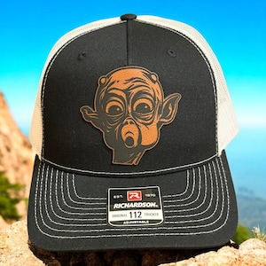 May include: Black and white trucker hat with a brown leather patch featuring a stylized alien face. The hat has a curved brim with white stitching and a white mesh back. The Richardson brand logo is visible.