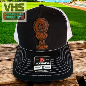 May include: Black and white trucker hat with a cartoon character patch. The hat features a curved brim, white mesh back, and the text "Richardson Original 112 Trucker Adjustable". The patch is a brown cartoon figure.