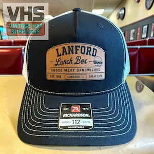 May include: A navy blue trucker hat with a white mesh back and a curved brim. The hat features a brown leather patch with the text "LANFORD Lunch Box" and a burger graphic. The brim has white stitching. The hat is adjustable.