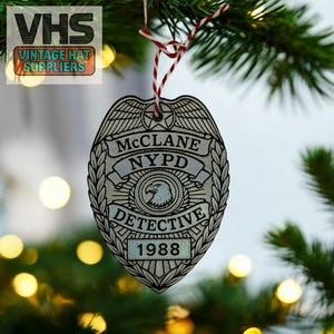 May include: A wooden Christmas ornament shaped like a police badge. The badge is silver and black with the text "McCLANE NYPD DETECTIVE 1988". The ornament hangs from a red and white string against a backdrop of a Christmas tree.