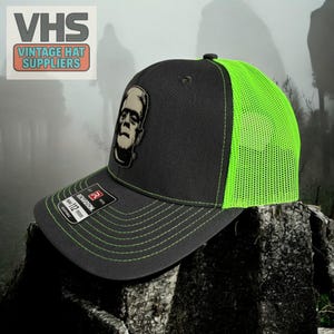 May include: Black and neon green trucker hat featuring a Frankenstein's monster patch. The hat has a curved brim, neon green mesh back, and adjustable snap closure. The hat has the text "VHS Vintage Hat Suppliers" on the upper left.