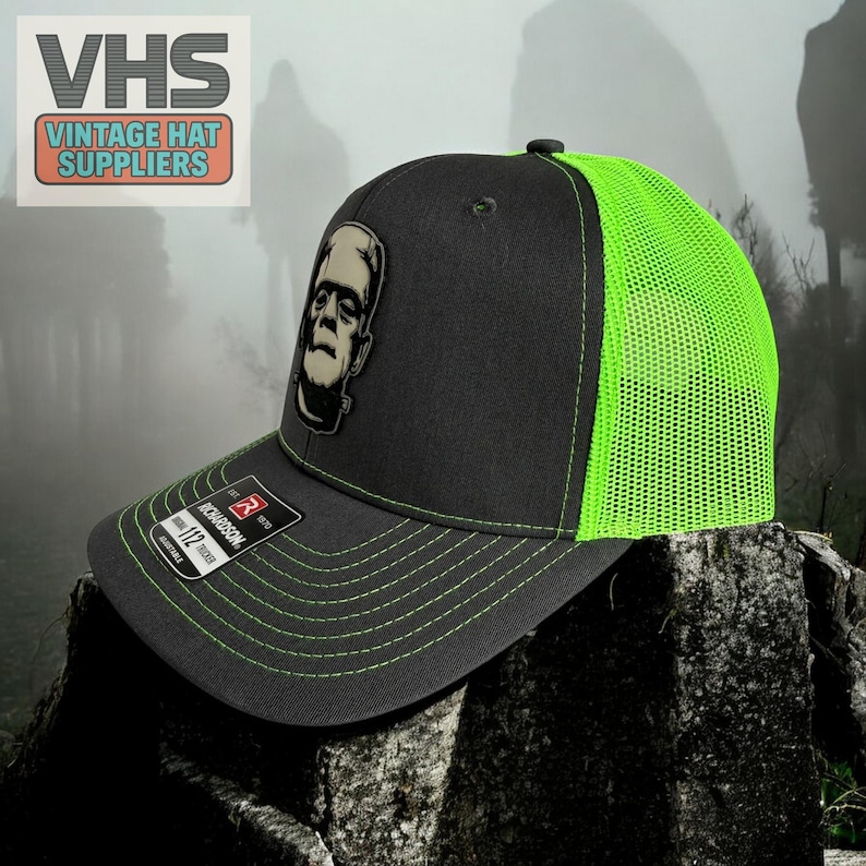 May include: Black and neon green trucker hat featuring a Frankenstein's monster patch. The hat has a curved brim, neon green mesh back, and adjustable snap closure. The hat has the text "VHS Vintage Hat Suppliers" on the upper left.