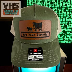 May include: Olive green trucker hat with a tan mesh back. The front features a brown leather patch with a covered wagon graphic and the text "You have typhoid." A Richardson 112 adjustable hat.