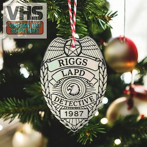 May include: A silver-colored Christmas ornament shaped like a police badge, hanging on a Christmas tree. The badge reads "RIGGS LAPD DETECTIVE 1987" and features an eagle emblem. The ornament is suspended by a red and white string.