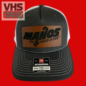 May include: A black and white trucker hat with a brown leather patch featuring the text "MANOS THE HANDS OF FATE". The hat has a curved brim and an adjustable strap. The hat is a Richardson Original 112 Trucker.