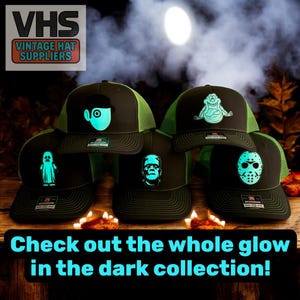 May include: A collection of black trucker hats with neon green mesh and glow-in-the-dark designs. The designs include a ghost, Frankenstein's monster, a monster, and a mask. The hats are displayed on a wooden surface with candles.