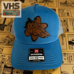 May include: A blue trucker hat with a cartoon graphic of a character with a rocket design. The hat has a mesh back and a curved brim. The hat has the text "RICHARDSON ORIGINAL 112 TRUCKER ADJUSTABLE". The hat also has the text "VHS VINTAGE HAT SUPPLIERS".