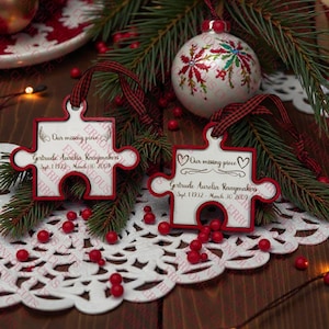Puede incluir: Dos adornos navideños en forma de rompecabezas con ribete y cinta rojos, con el texto "Our missing piece" y un nombre y fechas. Los adornos están rodeados de vegetación, bayas rojas y una bola navideña decorativa.