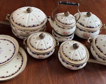 May include: A collection of cream-colored enamel cookware featuring a dark blue border with gold filigree and small pink floral accents. The set includes lidded pots, a tea kettle, and two flat plates, all topped with decorative metallic knobs.