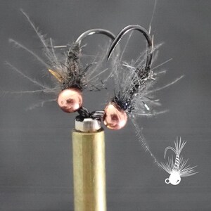 May include: Close-up of two fishing flies. Each fly features a copper-colored bead, black thread, and feathery accents. The hooks are silver, and the flies are mounted on a brass-colored base. A small white illustration of a fly is in the lower right corner.