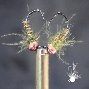 May include: Close-up of two fishing flies. Each fly features a curved hook, a body of green and brown fibers, and two copper-colored beads. The flies are mounted on a brass rod, with a white graphic of a fly in the lower right corner.