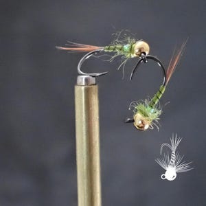 May include: Two handmade fishing flies. Each fly features a gold bead head, a curved hook, and a body of green and brown materials. The flies are designed for fly fishing. One fly is attached to a brass rod.