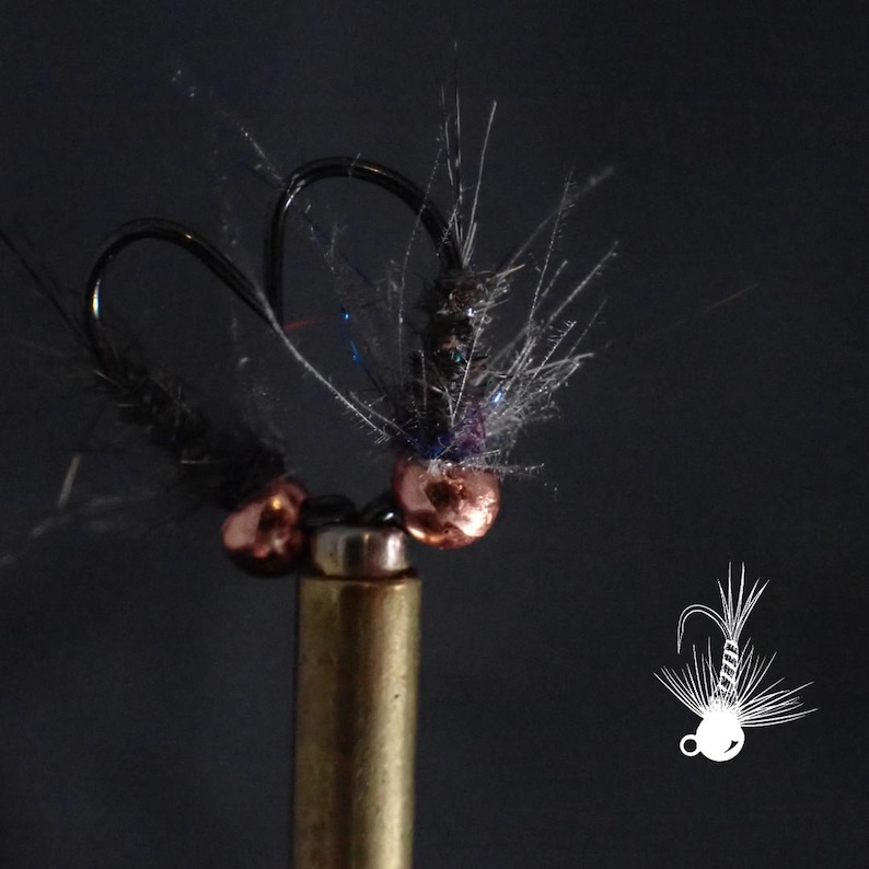 May include: Close-up of two black fishing hooks with copper-colored beads and dark gray and black feather-like accents. The hooks are mounted on a brass-colored base, set against a dark background. A white line drawing of a fishing lure is in the lower right corner.