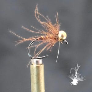 May include: Close-up of a handmade fishing fly. The fly features a gold-colored bead head, a dark hook, and brown, feathery materials. The fly is mounted on a brass-colored holder against a gray background. A white line extends from the hook.