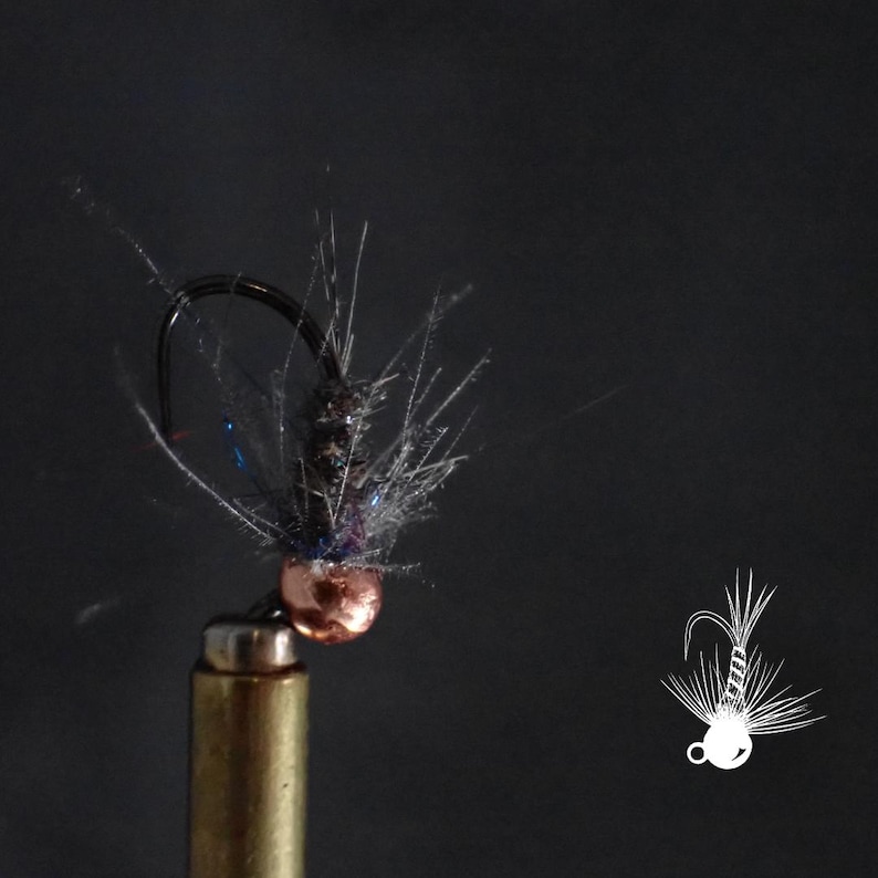 May include: Close-up of a fishing fly with a copper-colored bead head, black hook, and gray and blue feather accents. The fly is mounted on a brass-colored holder against a dark background. A white line drawing of a fly is in the lower right corner.