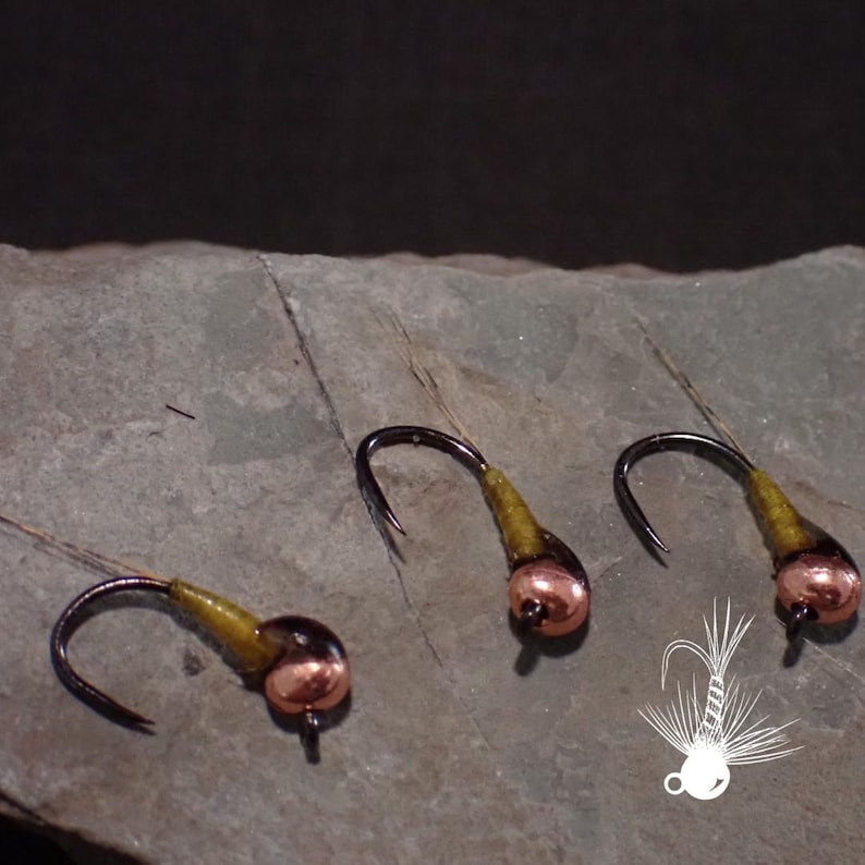 May include: Close-up of three fishing flies with copper-colored heads, black hooks, and olive-green bodies. The flies are arranged on a gray stone surface. A white graphic of a fly is in the lower right corner.