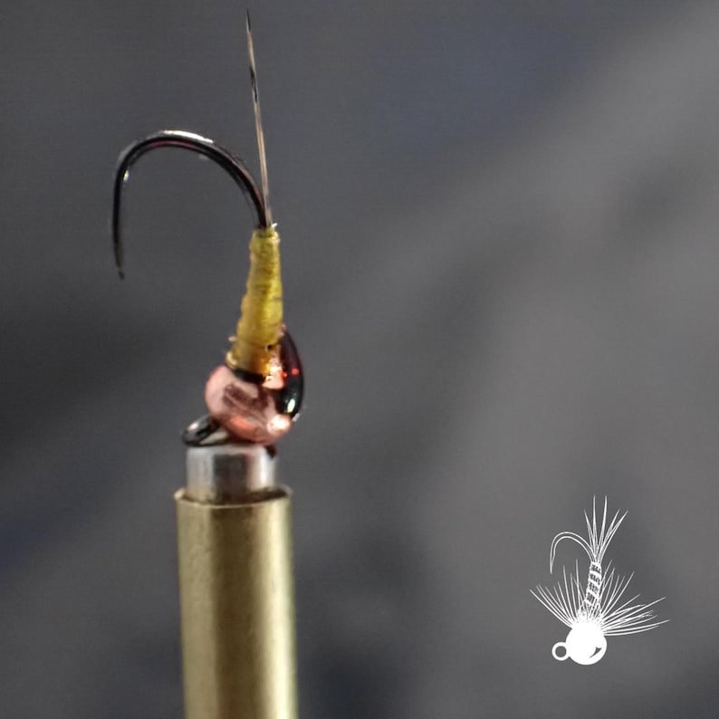 May include: Close-up of a fly fishing lure. The hook is black with a gold-colored body and a copper-colored head. A thin, silver-colored wire extends from the hook. The lure is mounted on a brass-colored stand.