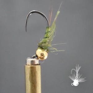 May include: A close-up of a fly fishing lure. The lure features a curved silver hook, a gold-colored bead head, and a body of green and tan materials. The fly is attached to a brass-colored cylindrical object.