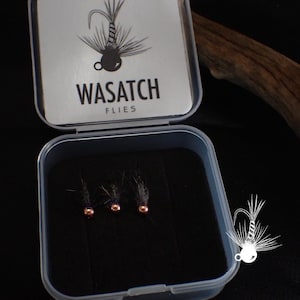May include: A small, open, clear plastic box containing three fishing flies. The flies have dark gray bodies and copper-colored beads. The box lid has the text "WASATCH FLIES" and a graphic of a fly.