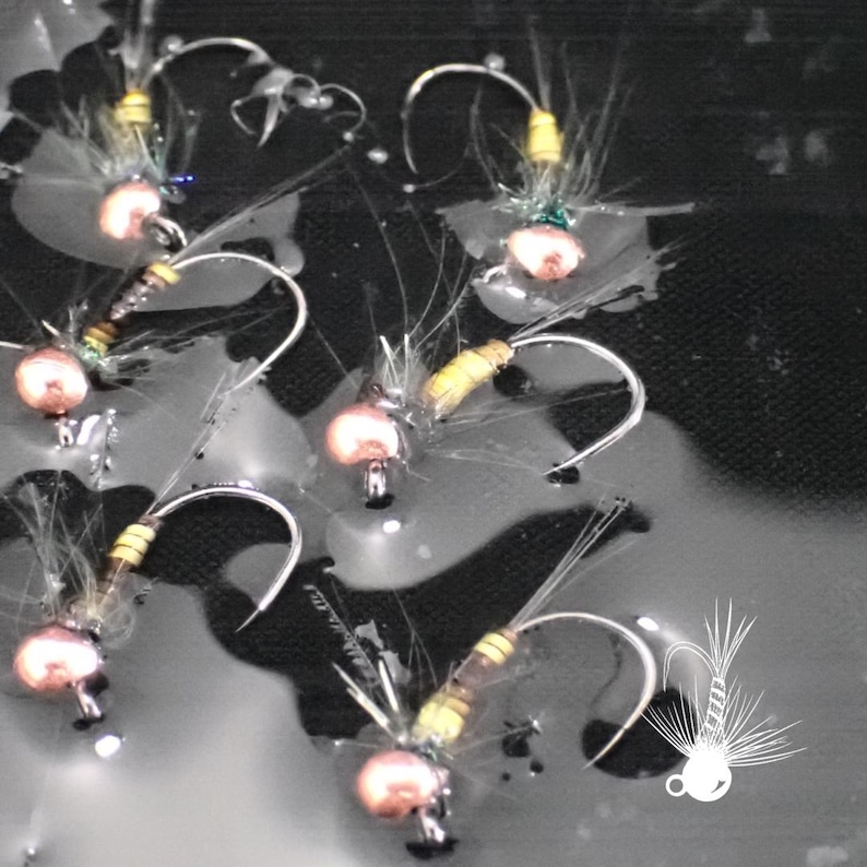 May include: Close-up of several handmade fishing flies. Each fly features a curved silver hook, a small, round, copper-colored bead, and various colorful feathers and threads. The flies are arranged on a dark, textured surface.