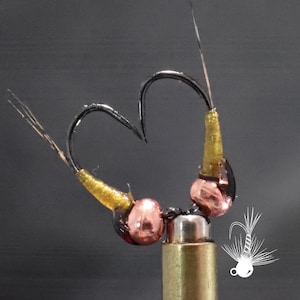 May include: Close-up of two fishing hooks arranged in a heart shape. The hooks are black with gold and copper-colored accents. The hooks are attached to a gold-colored base. The background is a dark gray.
