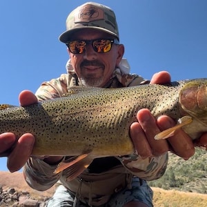 May include: A large rainbow trout with a speckled olive-green and silver body, held horizontally. The fish features a distinct pinkish-red lateral stripe along its side, a rounded snout, and translucent fins, showcasing its natural scales and texture.