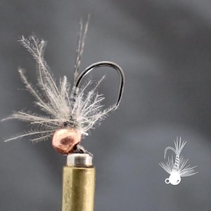 May include: Close-up of a fishing fly with a copper-colored bead head and a black hook. The fly features delicate, feathery white and gray materials, resembling wings. The fly is mounted on a brass-colored base against a gray background.