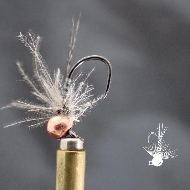 May include: Close-up of a fishing fly with a copper-colored bead head and a black hook. The fly features delicate, feathery white and gray materials, resembling wings. The fly is mounted on a brass-colored base against a gray background.