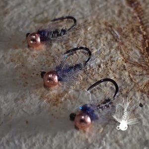 May include: Three fishing flies with copper-colored beads and black hooks. The flies have a fuzzy, blue and gray body. The flies are arranged on a textured, light brown surface. A small, white graphic of a fly is in the lower right corner.