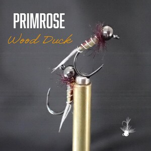 May include: A close-up of a fishing fly with the text "PRIMROSE Wood Duck". The fly has a silver hook, a silver bead, and a body with brown, white, and purple accents. The fly is mounted on a gold-colored stand.