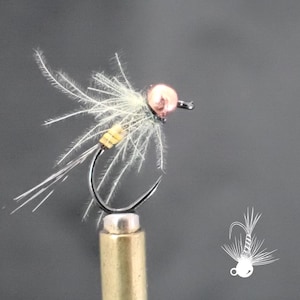 May include: Close-up of a fishing fly with a copper-colored bead head, a black hook, and white and yellow feather accents. The fly is designed to mimic an insect and is used to attract fish. The fly is mounted on a brass-colored base.