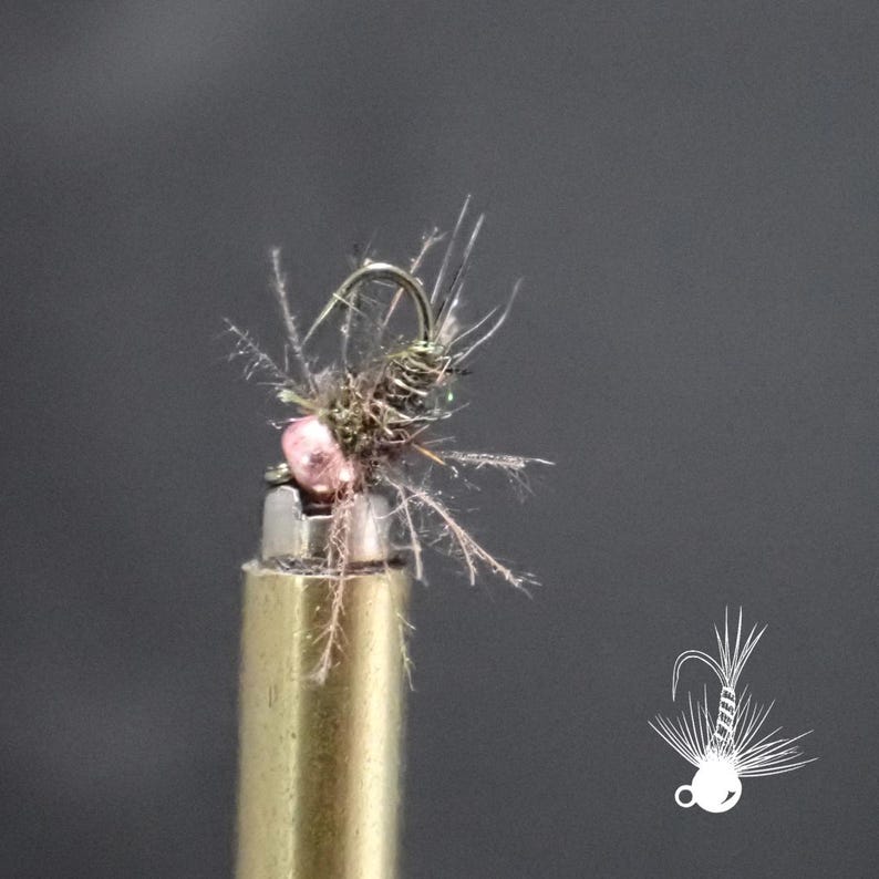 May include: A detailed image of a fishing fly. The fly has a curved hook, a pink bead, and a body made of brown and grey materials with feathery extensions. It is mounted on a brass-coloured object against a grey background.