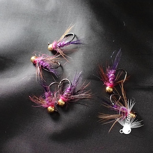 May include: Close-up of several fishing flies. Each fly features a gold-colored bead head, a body of purple and pink material, and brown and purple feather accents. The flies are arranged on a dark gray surface.