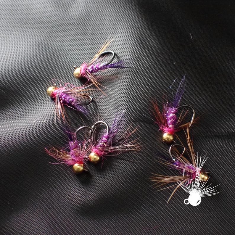 K&ouml;nnte beinhalten: Nahaufnahme von mehreren Angelk&ouml;dern. Jeder K&ouml;der hat einen goldfarbenen Perlenkopf, einen K&ouml;rper aus lila und rosa Material und braune und violette Federakzente. Die K&ouml;der sind auf einer dunkelgrauen Oberfl&auml;che angeordnet.