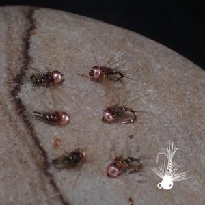 May include: Close-up of several artificial fishing flies. The flies have a metallic body, pink bead heads, and delicate, feathery wings. They are arranged on a light-coloured, textured stone surface. A white graphic of a fishing fly is in the lower right corner.