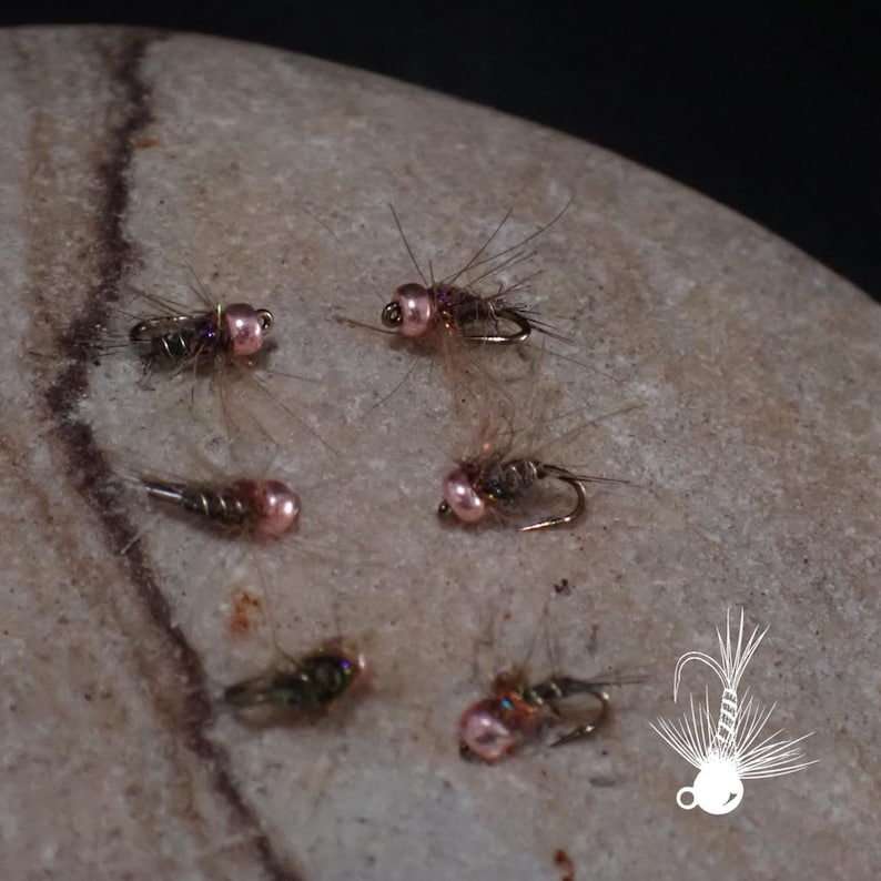 May include: Close-up of several artificial fishing flies. The flies have a metallic body, pink bead heads, and delicate, feathery wings. They are arranged on a light-coloured, textured stone surface. A white graphic of a fishing fly is in the lower right corner.