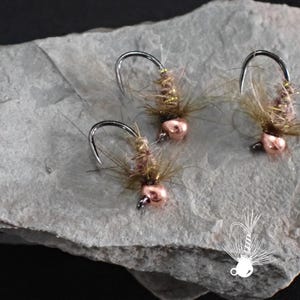 May include: Three fishing flies with copper-colored beads and curved hooks. The flies have a fuzzy, brown body with fine, dark fibers extending outward. They rest on a gray, textured stone surface, with a white logo in the bottom right corner.