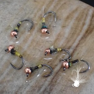 May include: Close-up of six fishing flies with curved hooks, copper-colored beads, and various colored thread and feather accents. The flies are arranged on a light brown surface, showcasing their intricate details.