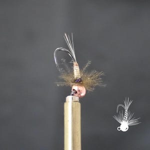 May include: A close-up of a handmade fishing fly. The fly features a silver hook, a tan body, and a cluster of brown and silver fibers. The fly is mounted on a gold-colored metal rod against a dark gray background.