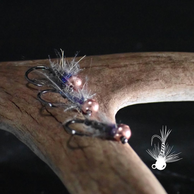 May include: Close-up of two fishing flies with gray and purple fibers, copper-colored beads, and curved hooks. The flies are resting on a piece of wood. A white graphic of a fishing hook is in the lower right corner.