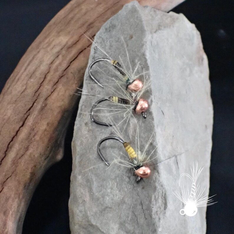 May include: Three fishing flies with curved hooks, copper-colored beads, and feathery white and tan fibers. The flies rest on a gray stone, with a piece of driftwood in the background. The flies are designed for fly fishing.