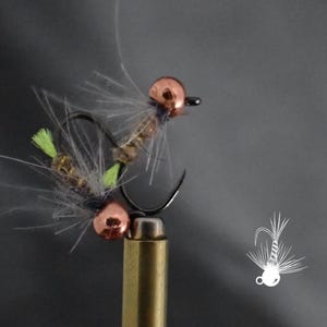 May include: Close-up of two fishing flies. Each fly features a copper-colored bead head, a black hook, and a body with gray, green, and brown elements. The flies are designed for fly fishing.