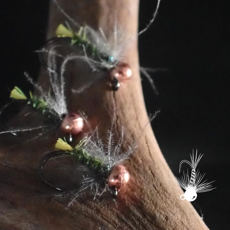 May include: Close-up of three fishing flies. Each fly features a metallic pink bead, a dark hook, and a combination of green, yellow, and white feathers. The flies are arranged on a piece of wood against a dark background.