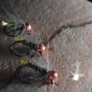 May include: Three fishing flies with curved hooks, copper-colored heads, and green bodies with yellow accents. The flies are arranged on a textured, light-colored stone surface. The flies are designed to mimic insects.