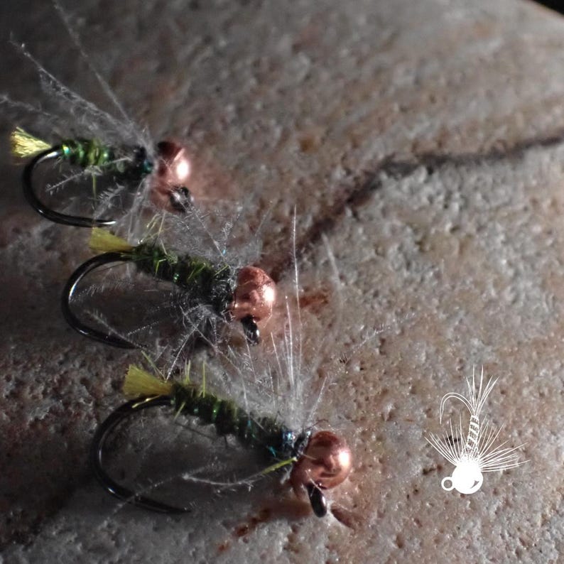 May include: Three fishing flies with curved hooks, copper-colored heads, and green bodies with yellow accents. The flies are arranged on a textured, light-colored stone surface. The flies are designed to mimic insects.