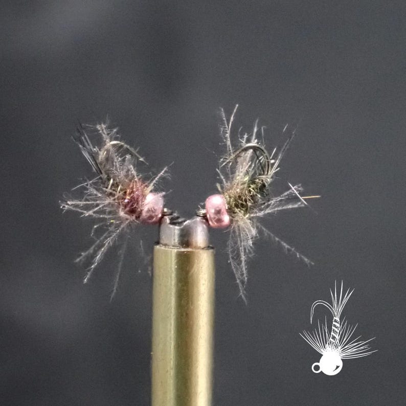 May include: Two handmade fishing flies with pink beads and feathery details are mounted on a brass rod. The flies have a complex design with fine, light-coloured fibres and dark hooks, set against a dark grey background.